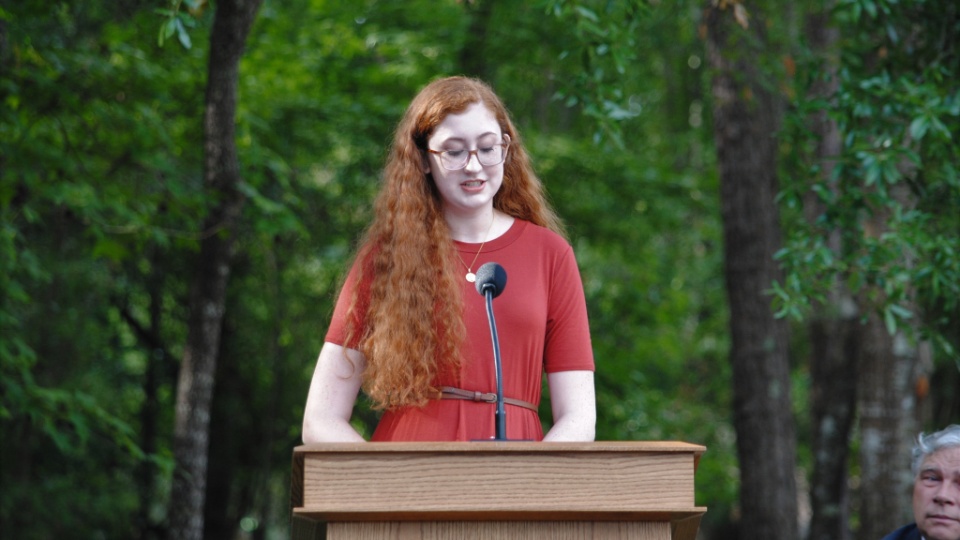 A woman in a red dress and glasses speaking from a pulpit outside.