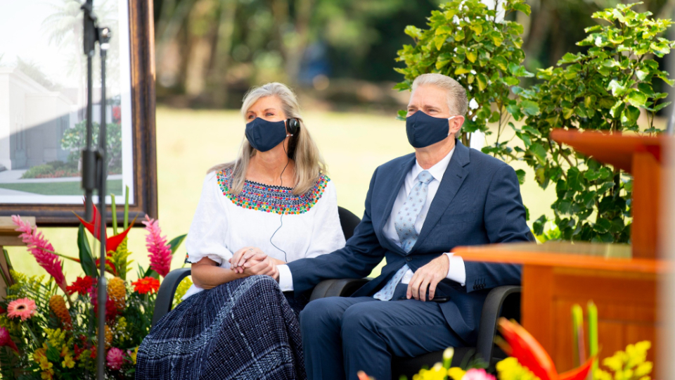 A man in a suit and a woman in a dress sitting in chairs outside, holding hands and wearing face masks.
