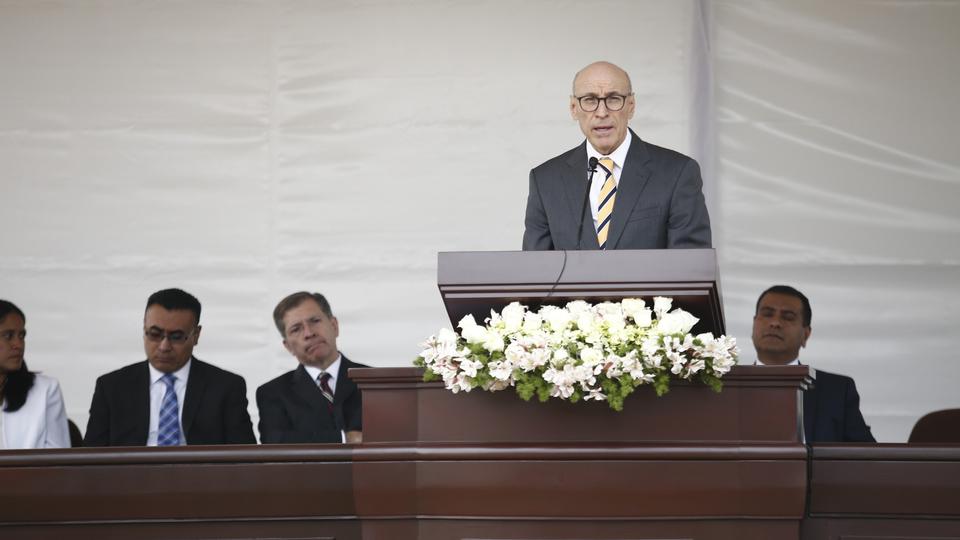 A man in glasses, a suit coat and a yellow tie speaking at a pulpit outside. 