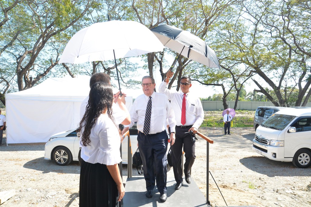 Elder Jeffrey R. Holland arriving at the Urdaneta Philippines Temple site, with two Latter-day Saints next to him holding umbrellas to block sunlight.