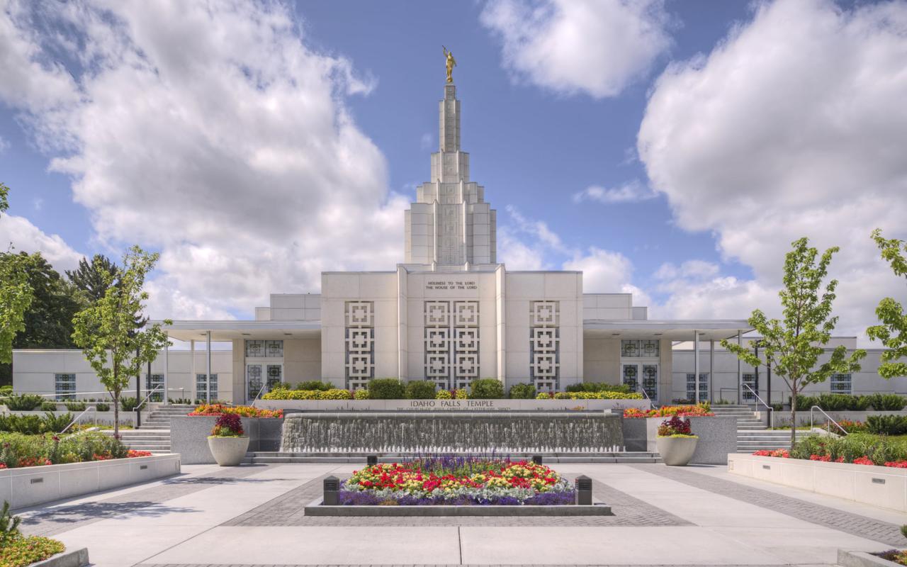 The Idaho Falls Idaho Temple, a white, multilevel building, with trees and a river in the foreground.