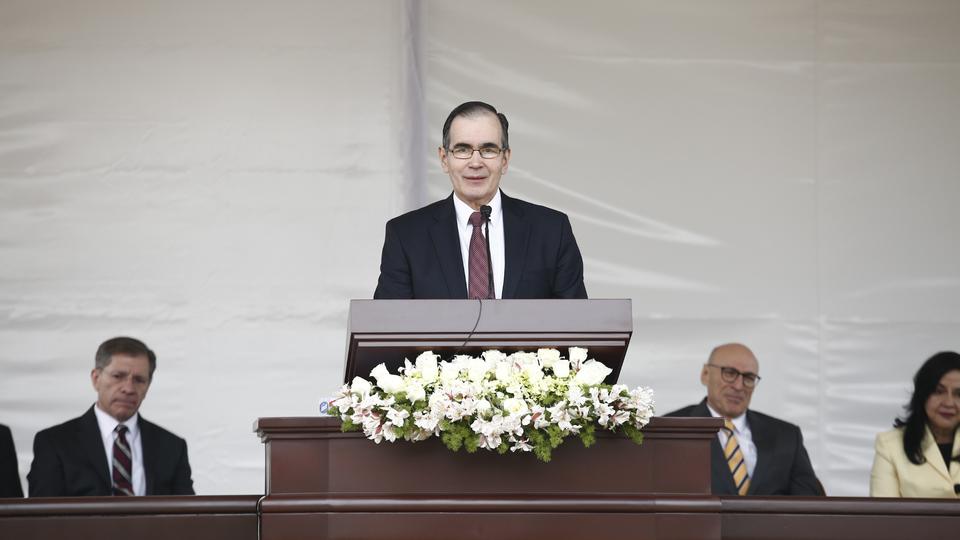 A man wearing glasses, a suit and a tie, speaking from a pulpit outside.