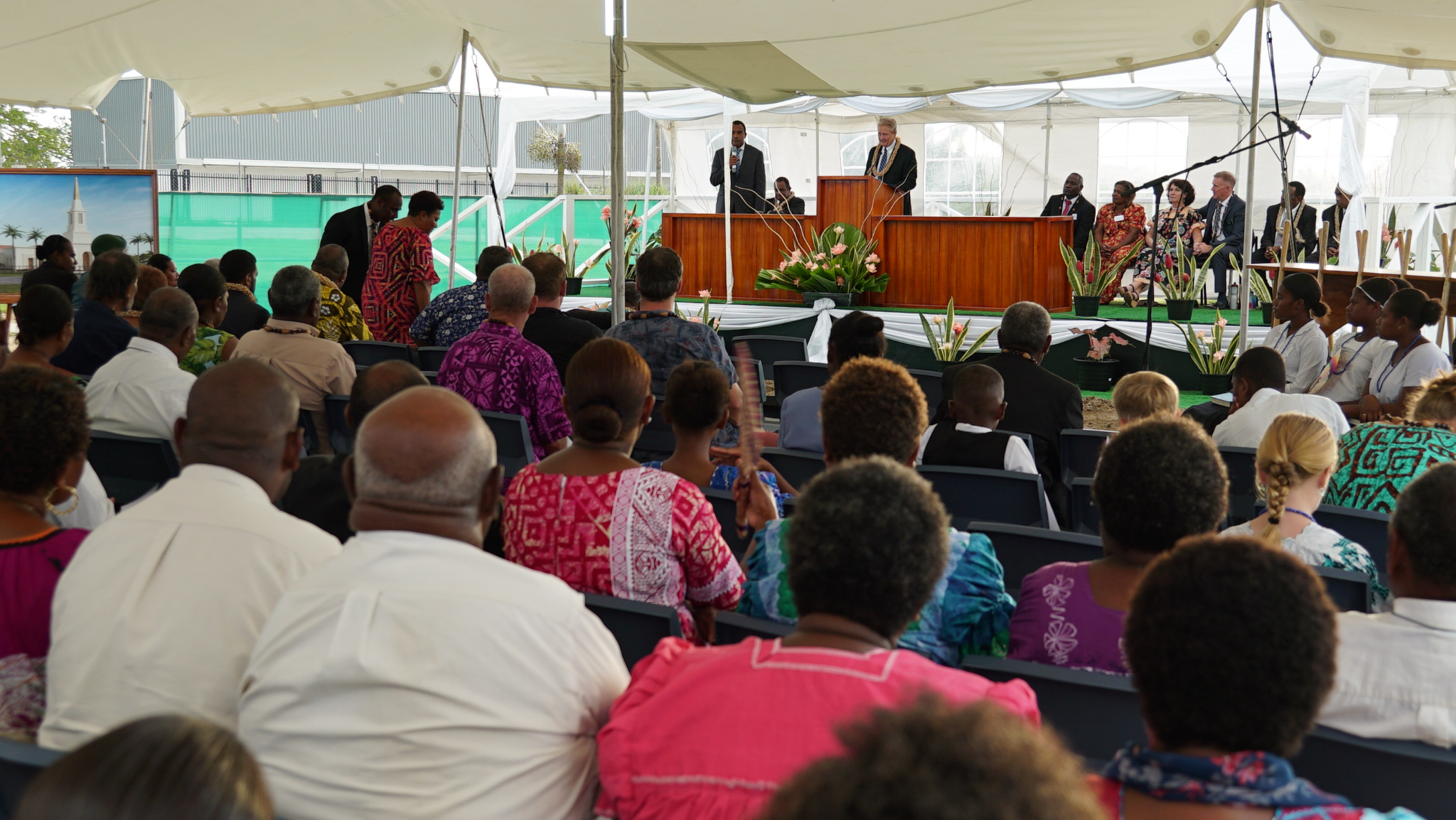 A large congregation of people under a cloth shade structure outside, listening to a man in a suit speak at a pulpit.
