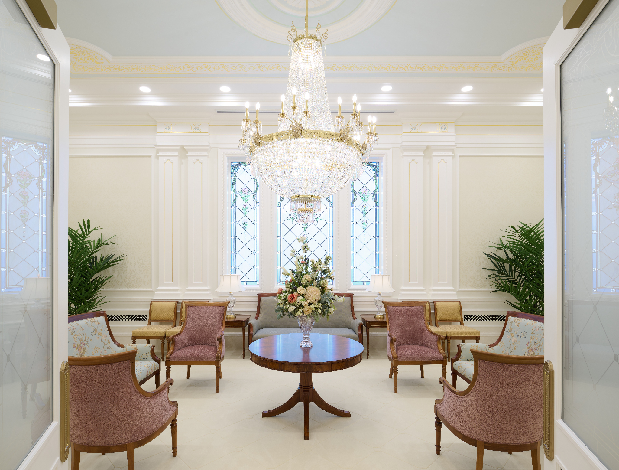 A white room with a clear chandelier in the center, rectangular windows on one wall, and dark-wood chairs around the room. 