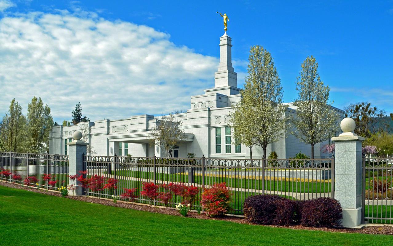 The Medford Oregon Temple, a white one-story building with skinny, rectangular windows and a center multilevel spire with the angel Moroni. A fence is also seen around the temple grounds, with red bushes outside of the fence.
