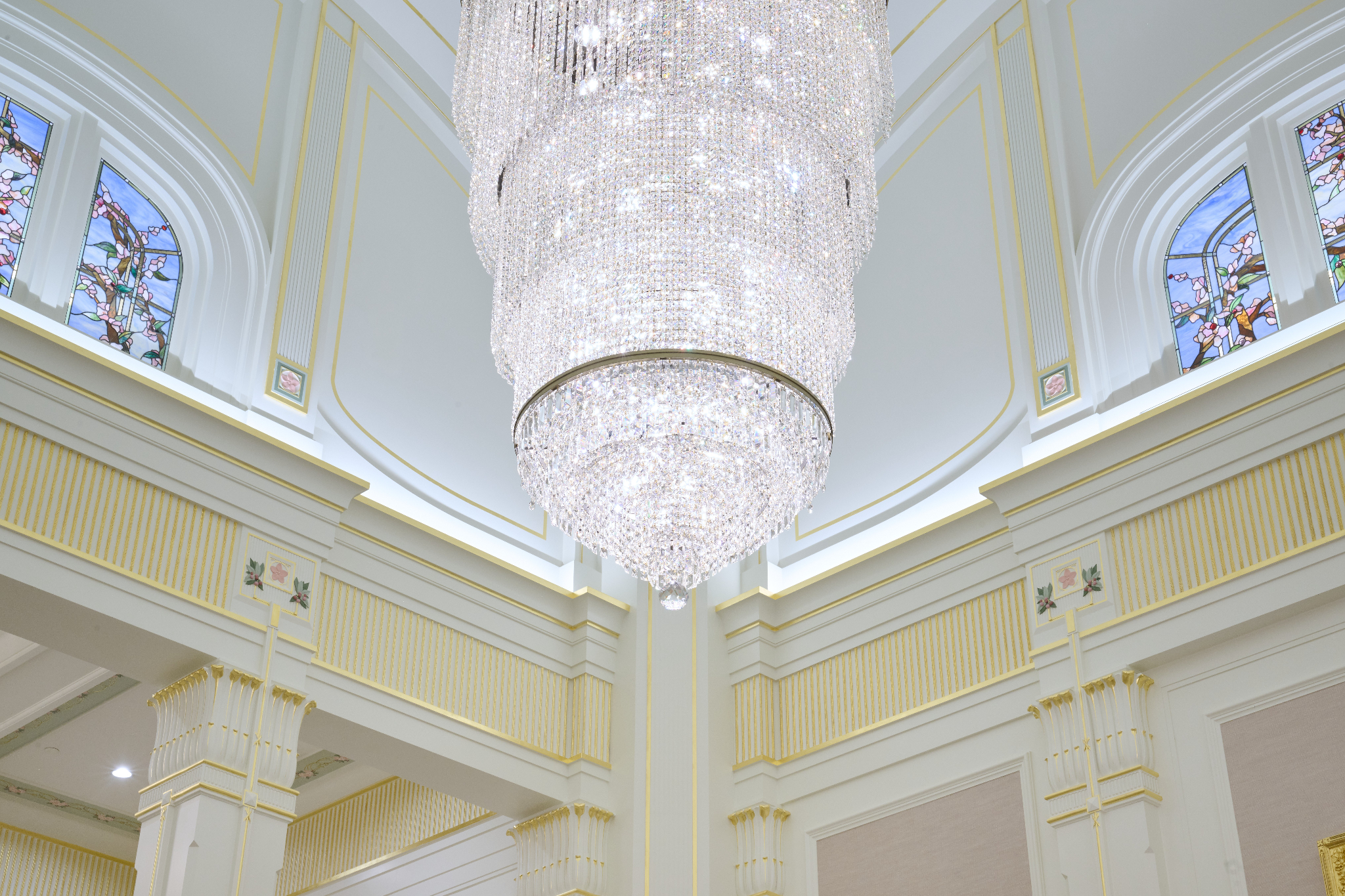 A large, clear chandelier hanging from a tall ceiling in a white room.