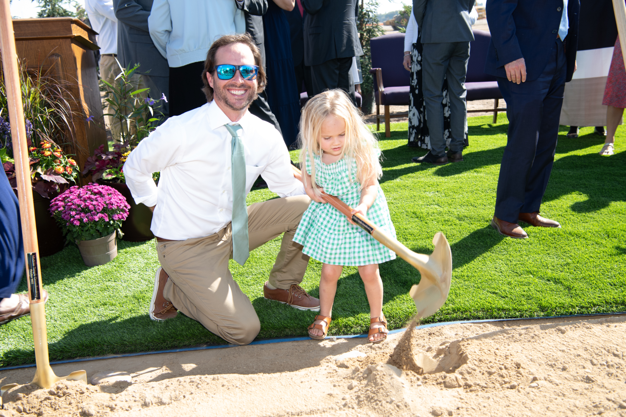 A dad kneeling next to his child daughter, with the daughter holding a shovelful of dirt.