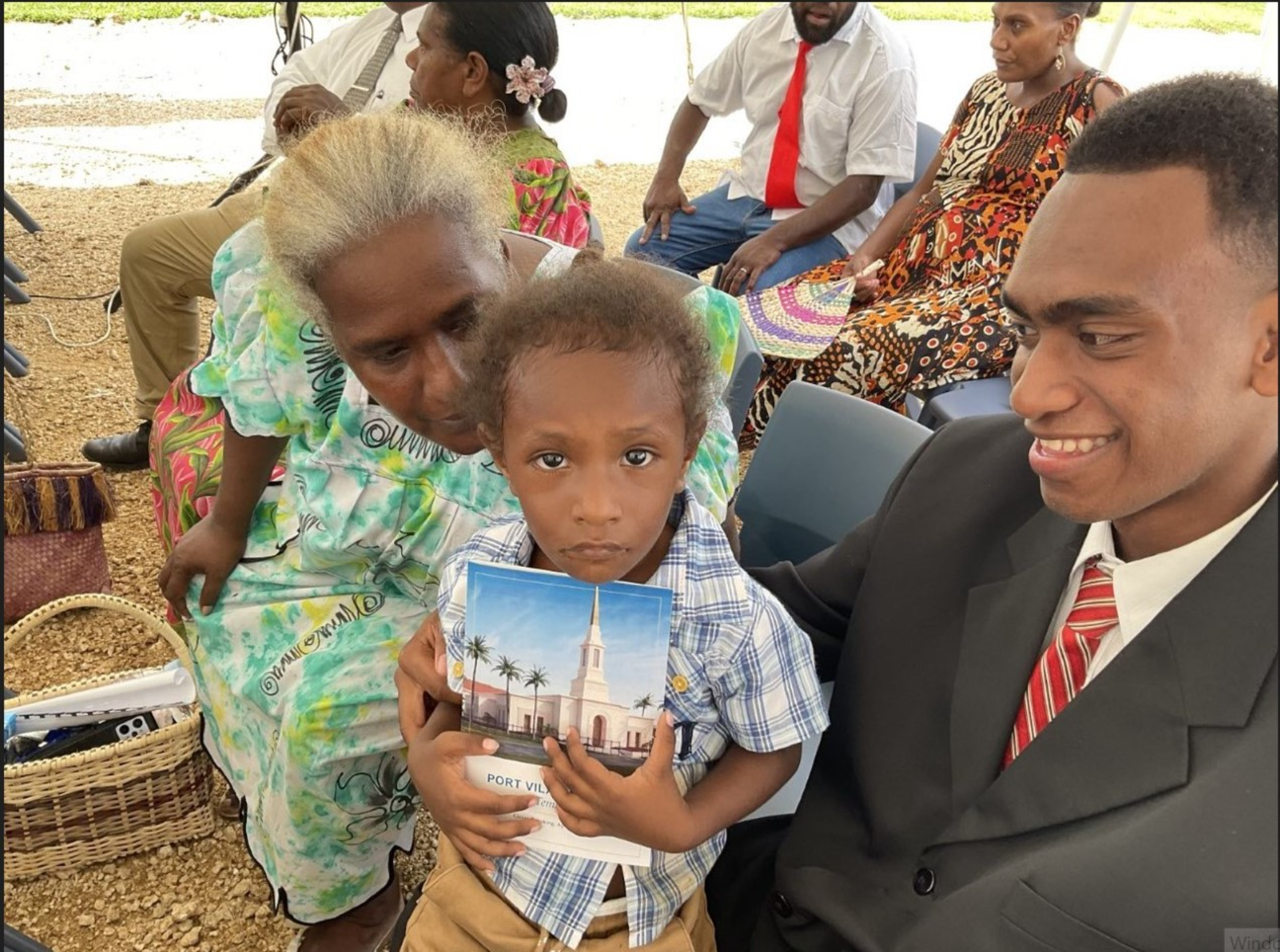 A boy holding a paper program that shows a rendering of the Port Vila temple, with several other people in Sunday best seated around him.