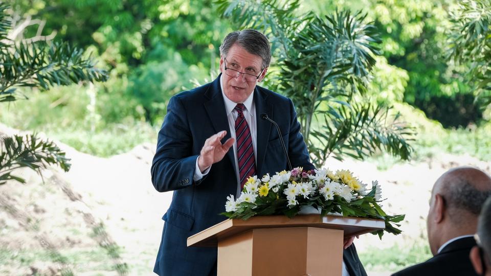 A man wearing a suit, tie and glasses and speaking from a pulpit outside, with trees in the distance behind him.