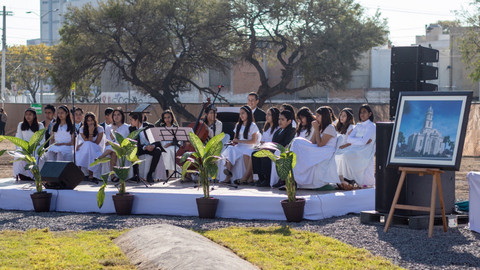 A choir of young women wearing white dresses and young men wearing black suits, all sitting on a long, white platform.