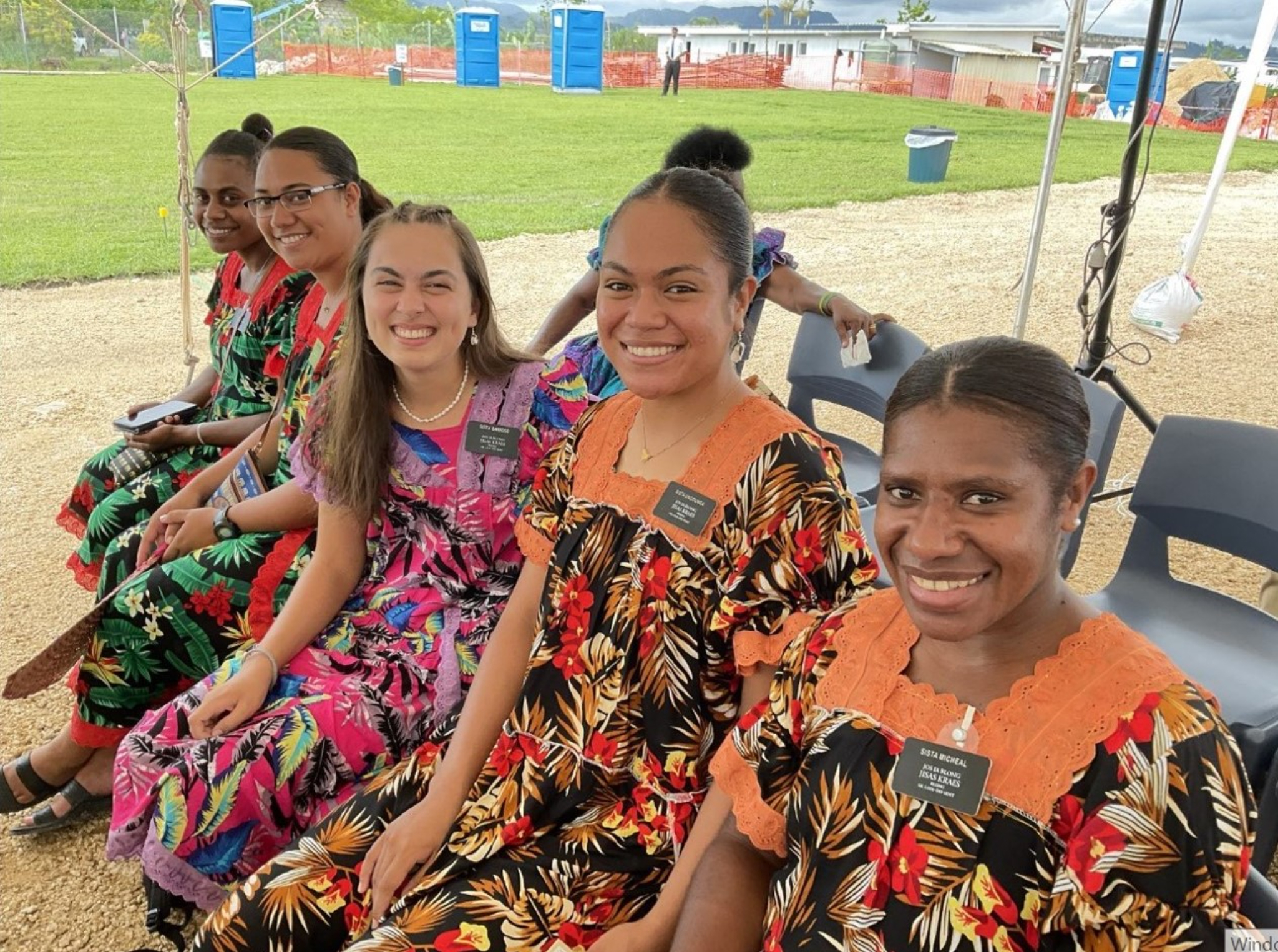 A row of five sister missionaries wearing colorful flower-patterned dresses and smiling.