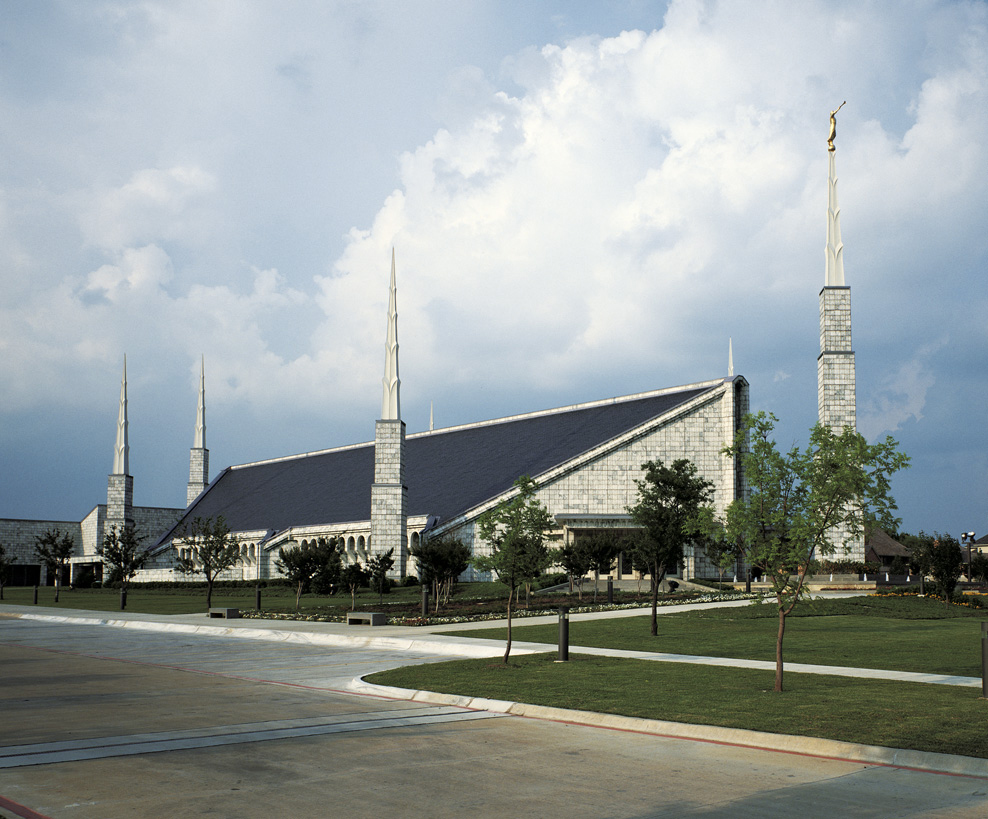 The exterior of the Dallas Texas Temple, a building with a peaked roof and six detached spires standing around the edifice.