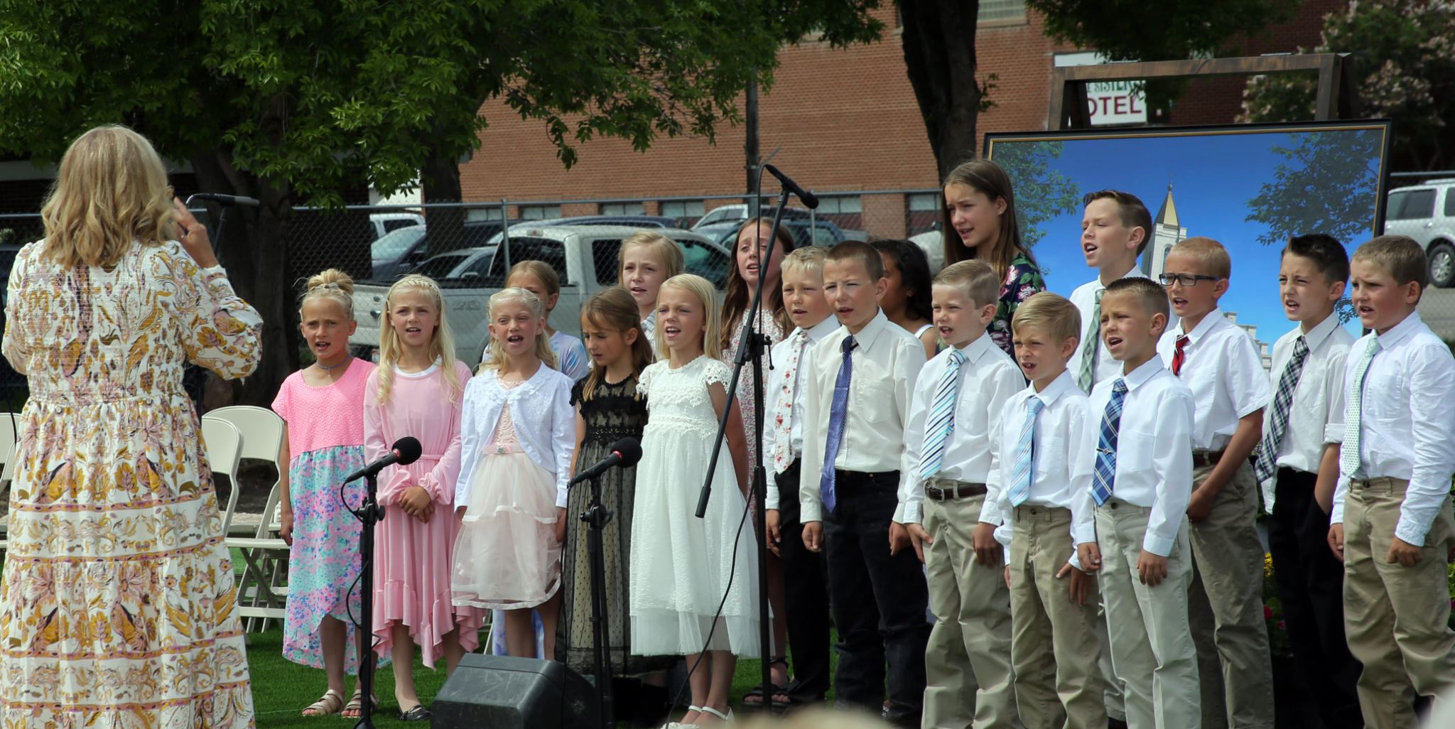 A group of Primary-aged boys and girls in Sunday best singing, with a woman in a flowery dress conducting them.