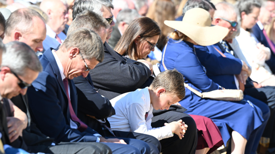A crowd of people sitting in chairs outside and bowing their heads in prayer.