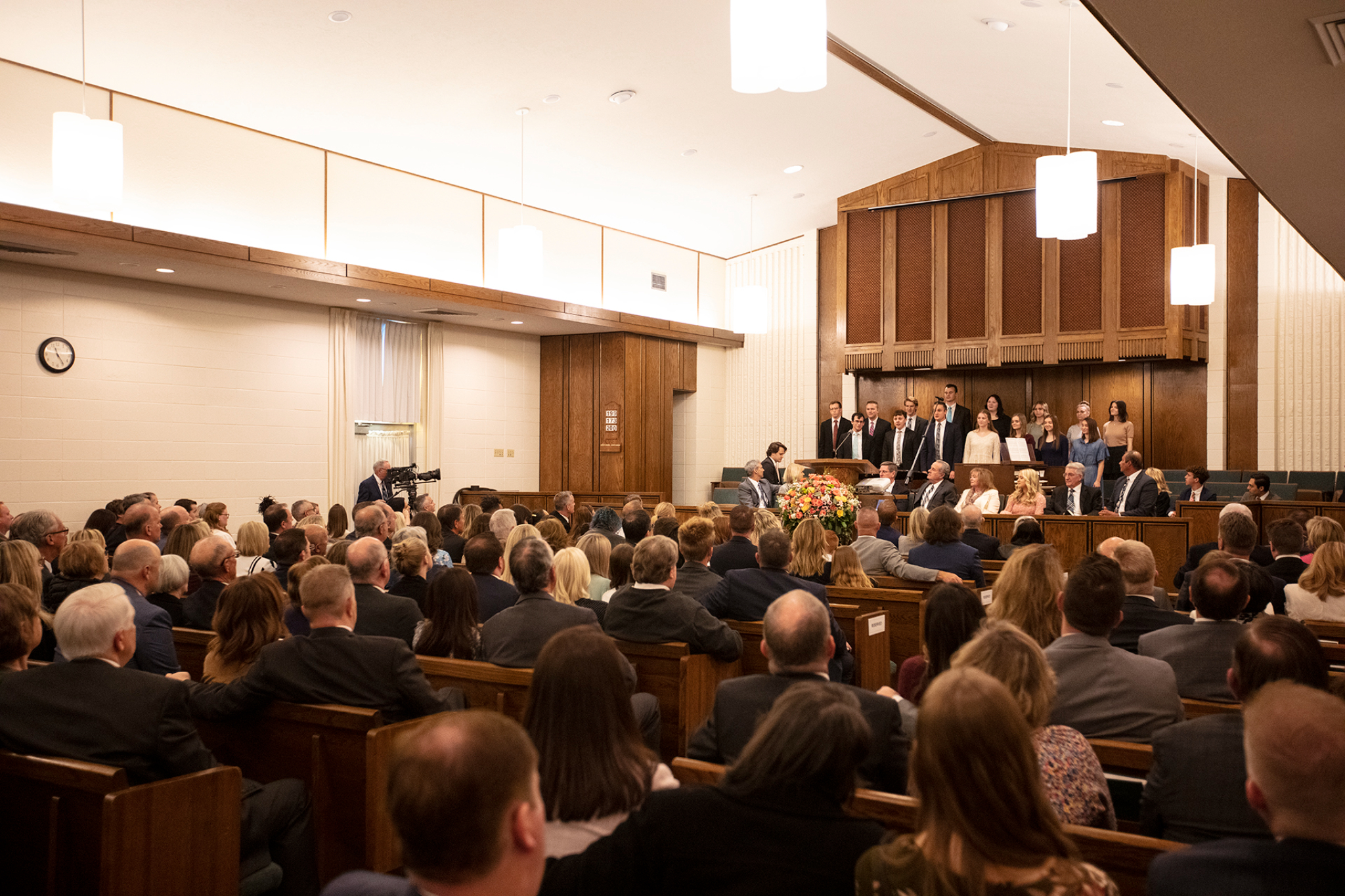 A congregation inside a Latter-day Saint chapel, with a choir of men in suits and women in dresses at the front of the room.