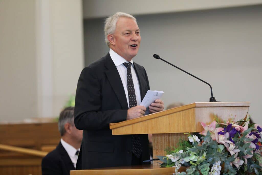 A man wearing a suit and tie and speaking from a pulpit indoors.