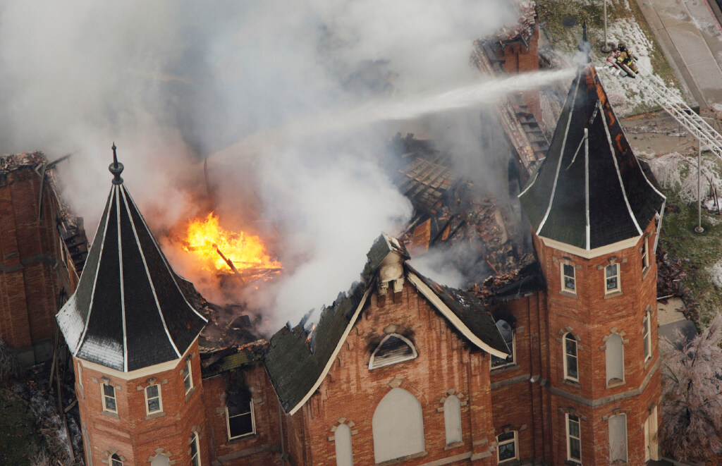 An aerial view of the Provo City Center Temple, a red-brick building, with fire and smoke coming from the top and streams of water pointed at the building.