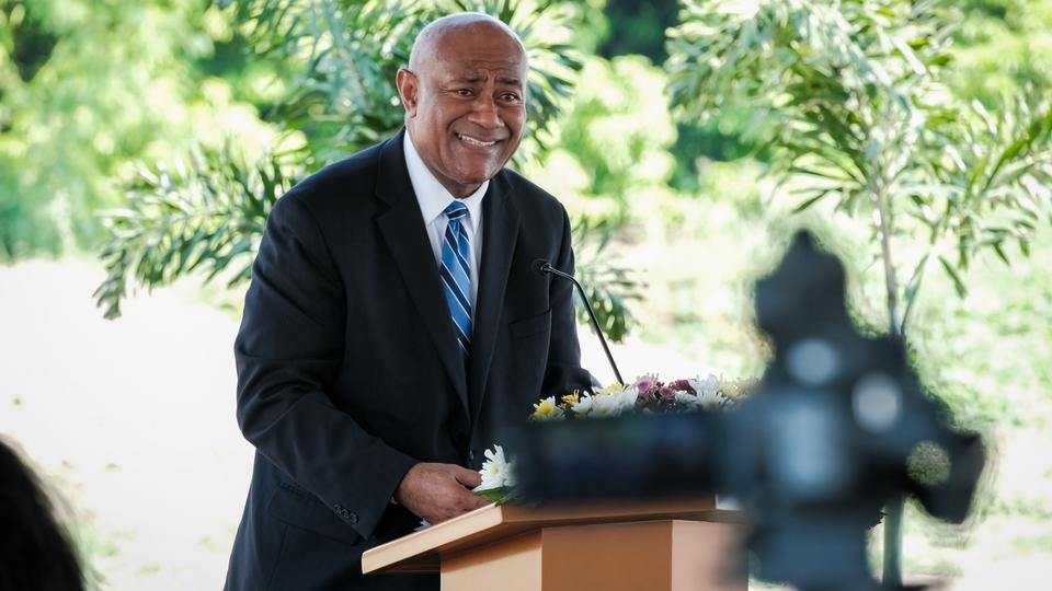 A man wearing a suit and tie smiling and speaking from a pulpit outside.