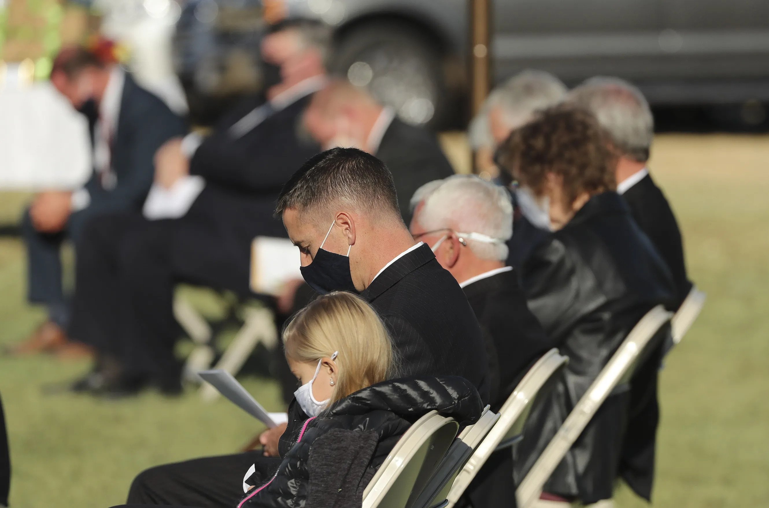 A row of people in Sunday best, sitting on chairs outside and bowing their heads in prayer.