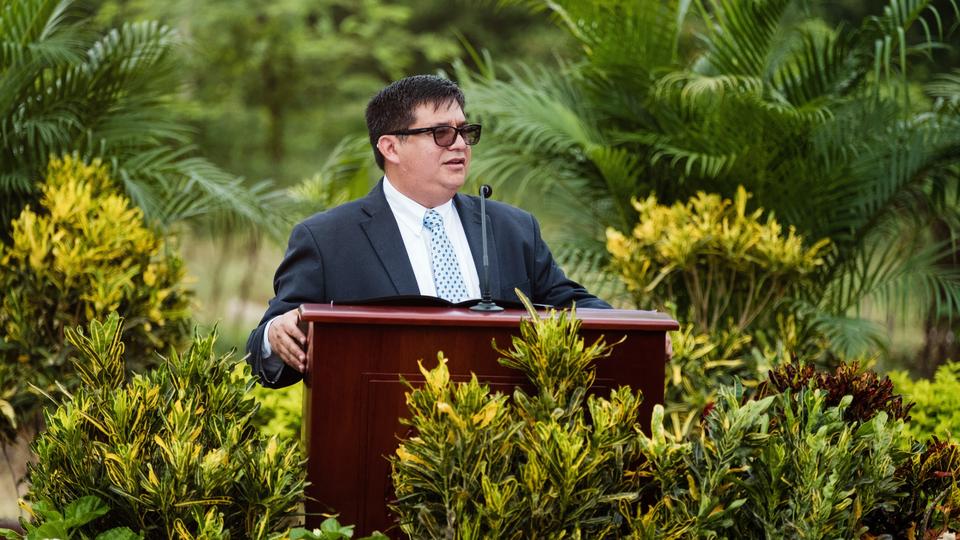 A man wearing a suit, tie and glasses and speaking from a pulpit outside with a forest behind him.