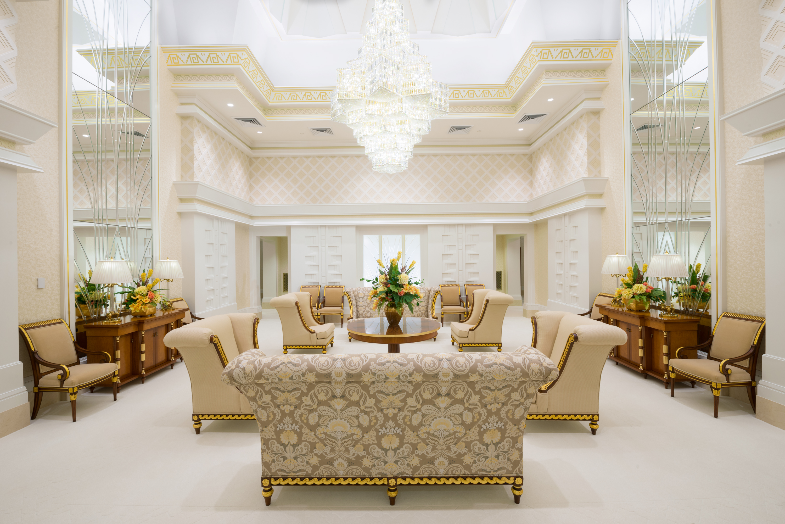 An open white room with armchairs, couches with flower designs, large mirrors and a large clear chandelier.