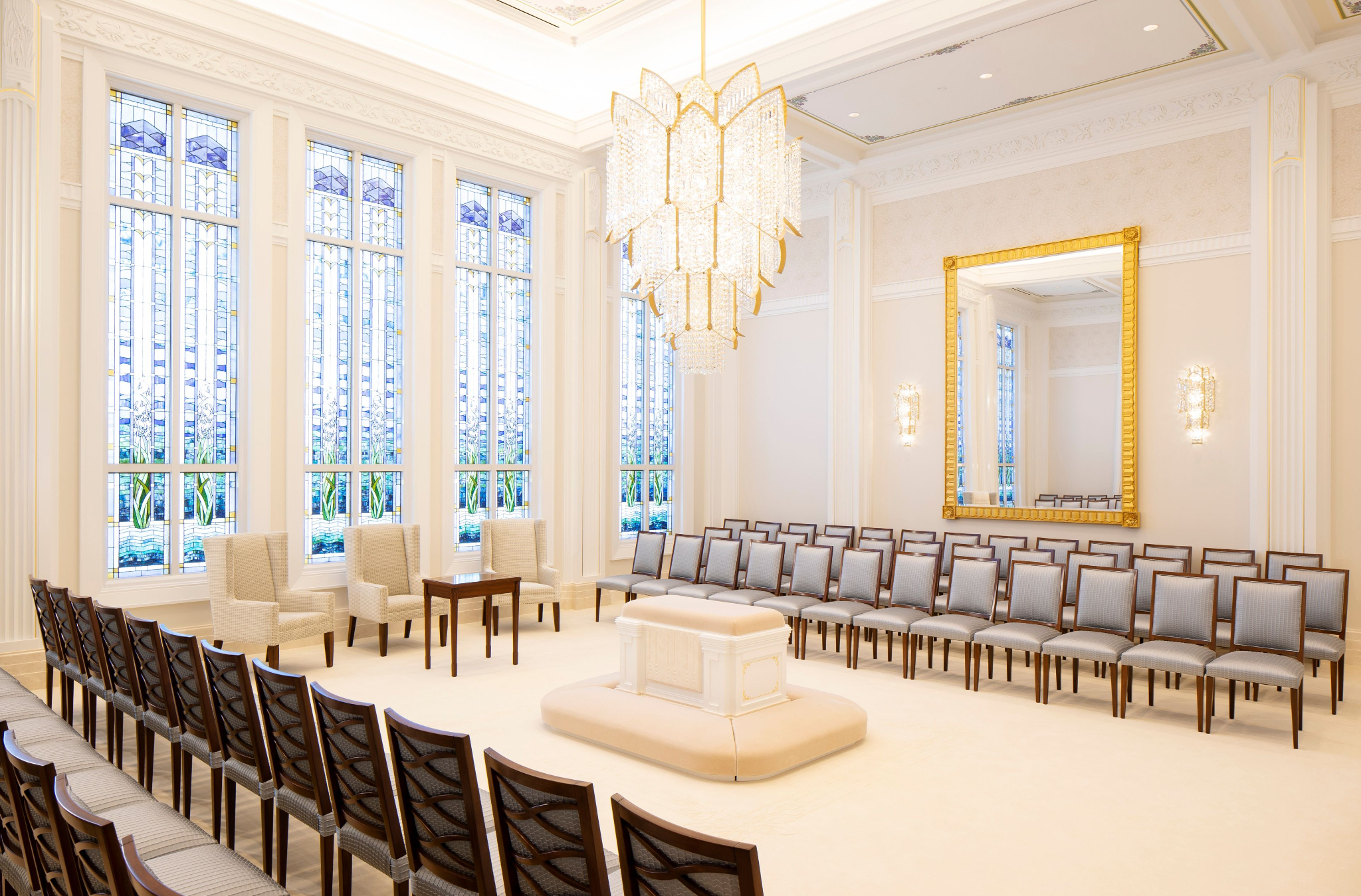 A white room with rows of chairs on two sides, a white altar in the center, and a rectangular mirror with a golden border on the wall.