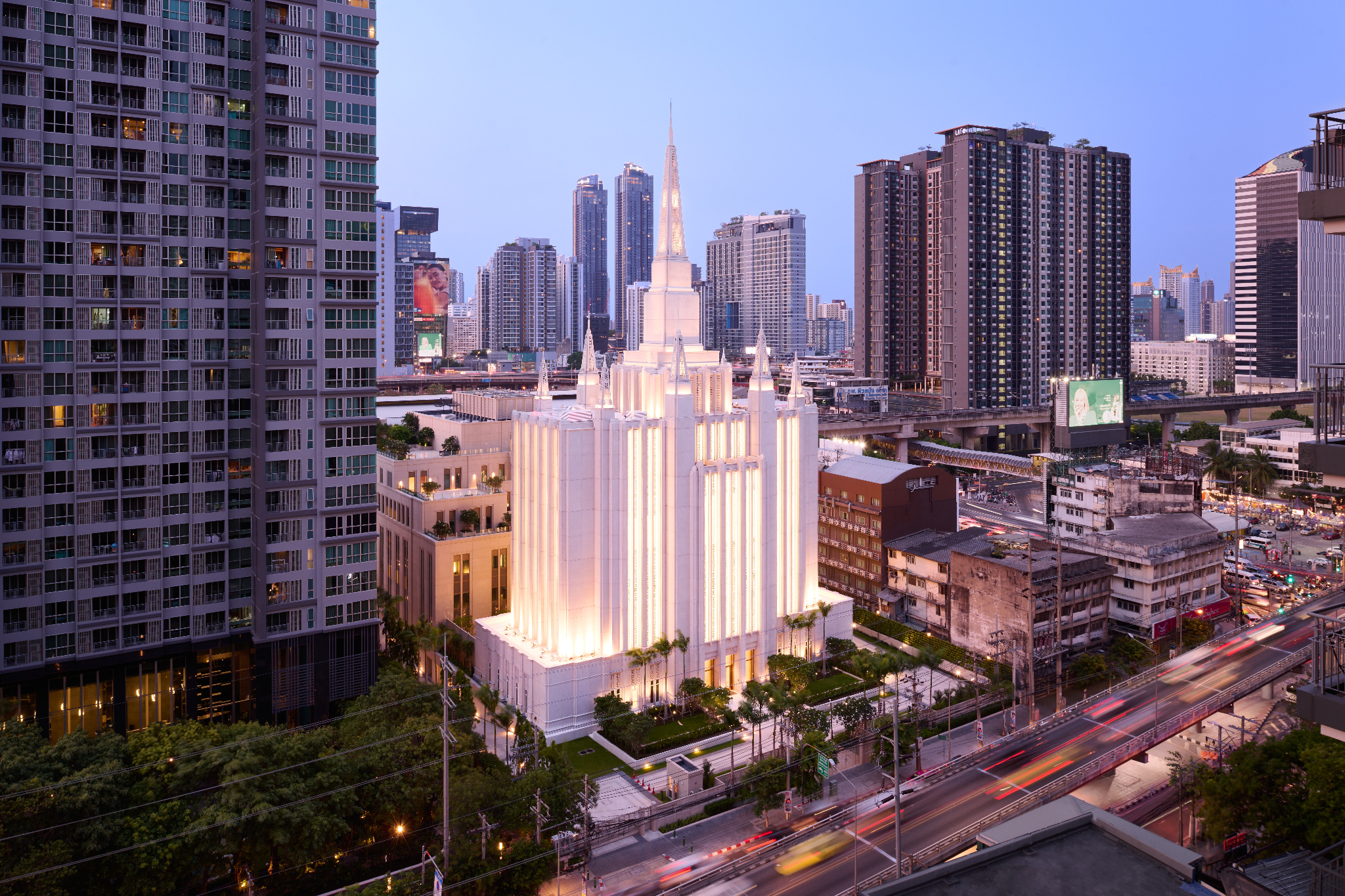 The exterior of the Bangkok Thailand Temple, a tall, off-white building with small spires on top and several skyscrapers around it. 