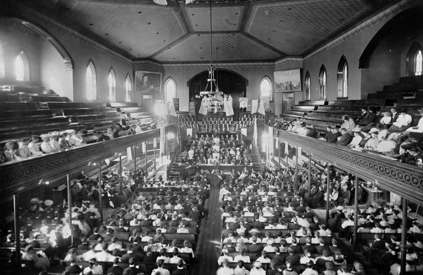 An open room with a large congregation of people sitting in benches.