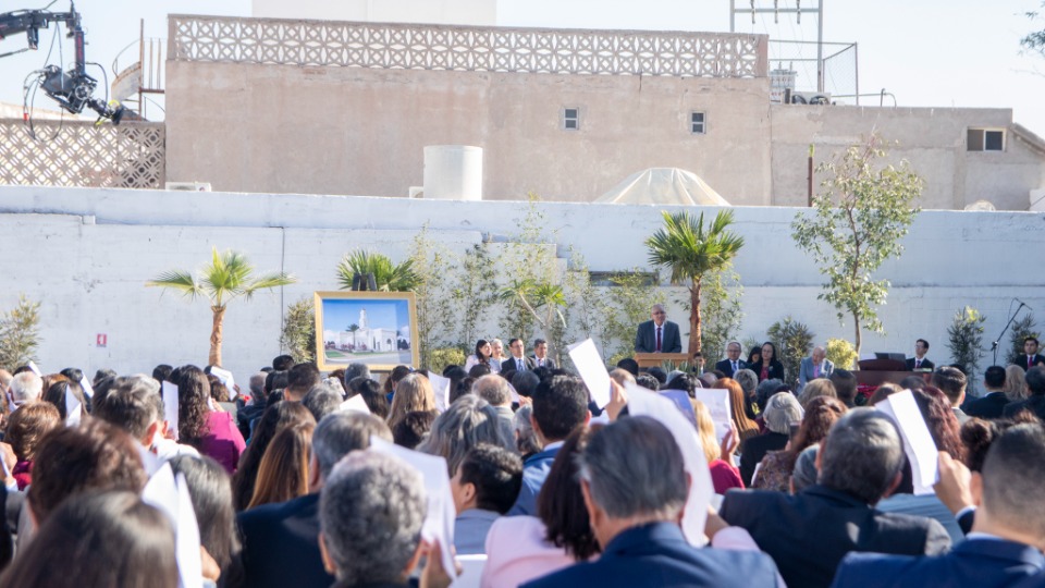 A crowd of people sitting outside on white chairs at the Torreón Mexico Temple groundbreaking ceremony while listening to Elder Hugo Montoya speak from a pulpit.