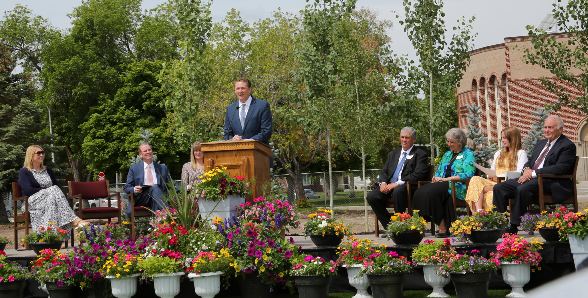 Elder Ryan K. Olsen, a man wearing a suit and tie, speaking from a pulpit outside, with many flower pots on the ground around him.