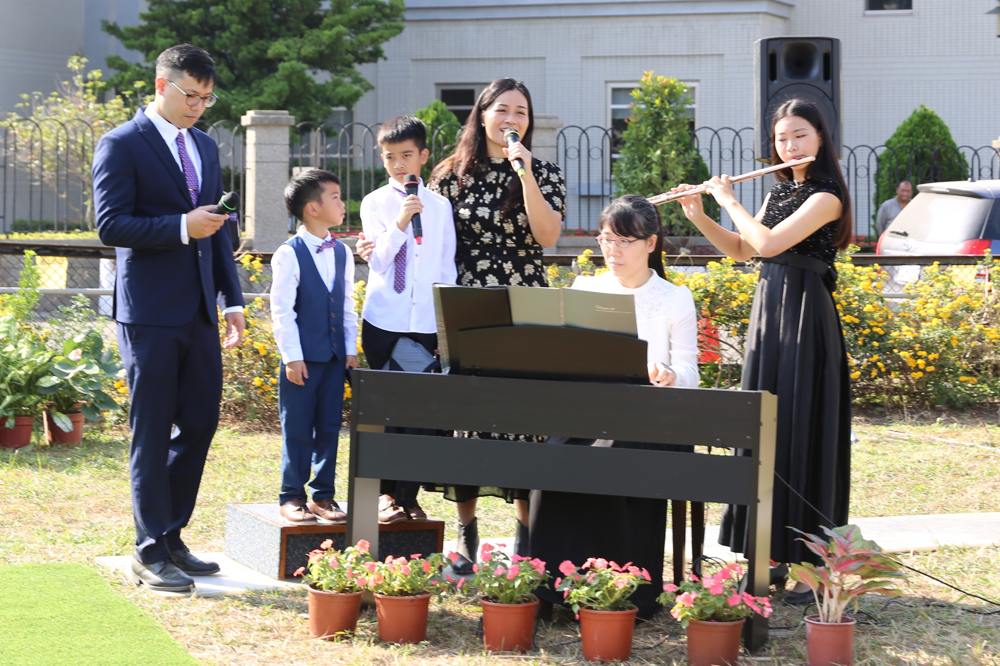 A man, a woman and two boys singing next to a woman playing piano and a young woman playing the flute.