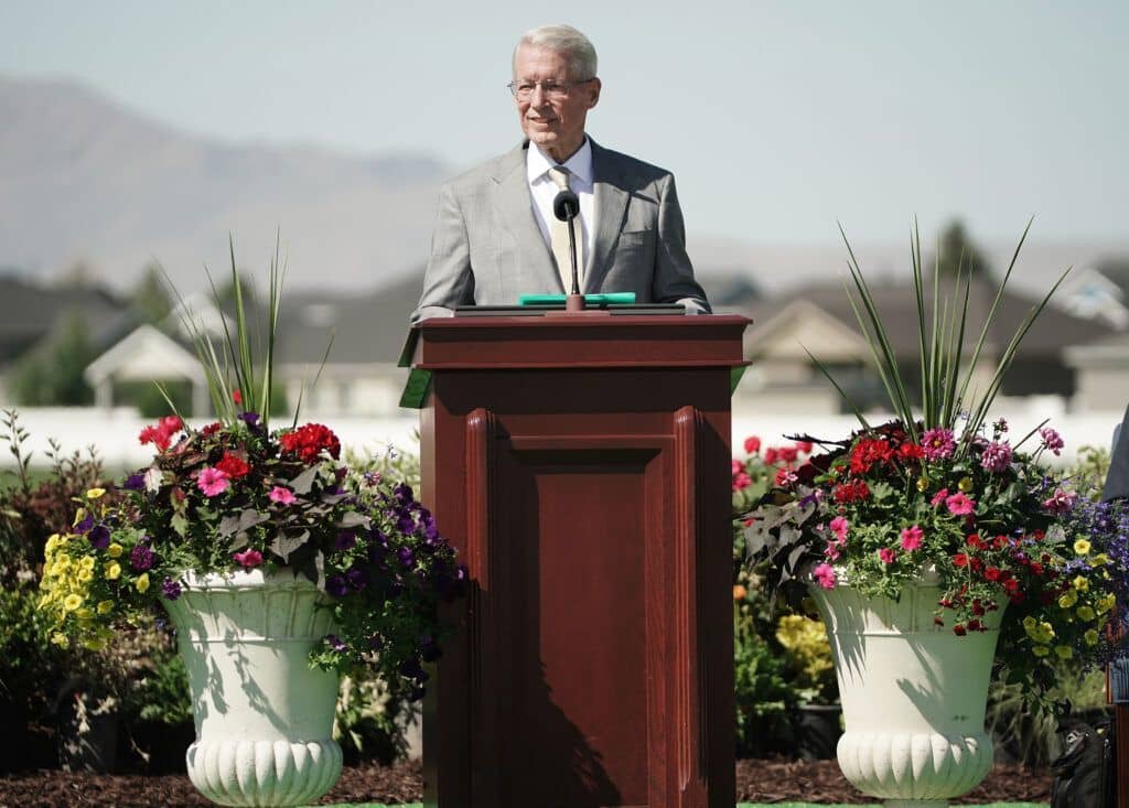 A man wearing a suit, tie and glasses and speaking from a pulpit outside.