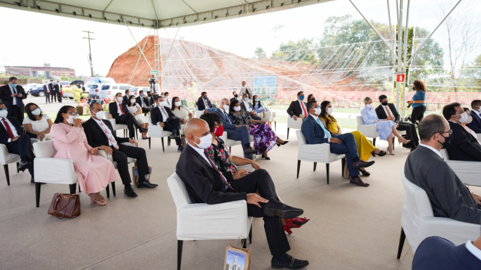 A congregation of people wearing Sunday best and face masks sitting in chairs outside under a shade structure.