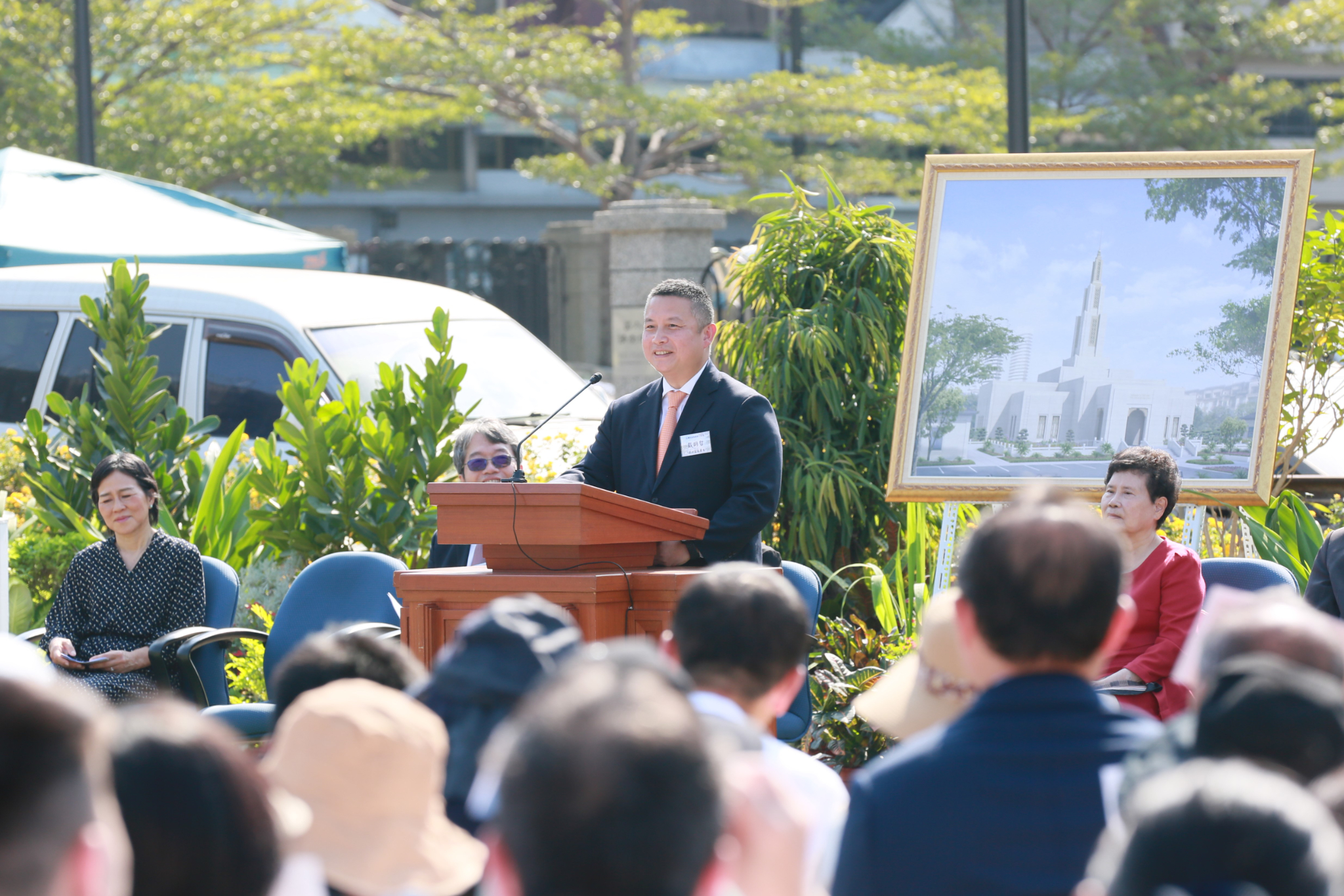 Elder Benjamin M. Z. Tai wearing a suit and tie and speaking from a pulpit outside.