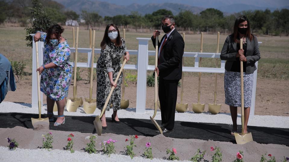 Three women in dresses and one man in a suit holding ceremonial golden shovels planted into the ground. 