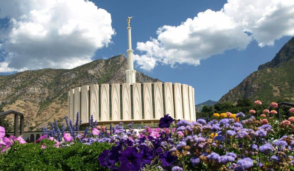 The Provo Utah Temple, a flat white building with a thin spire on top, from a low angle with flowers all around.