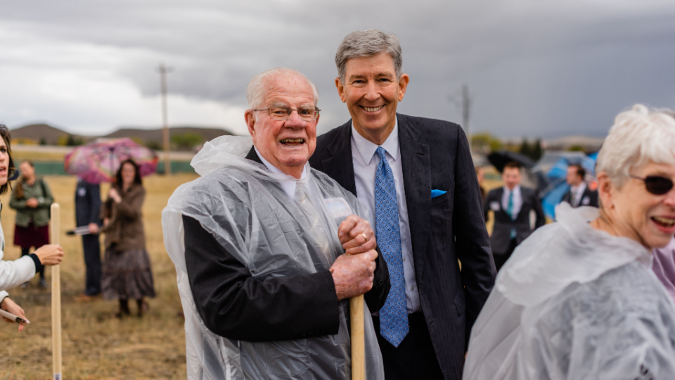 Two men in suits smiling at the camera, one of the men with a rain poncho, with several people with umbrellas behind them.