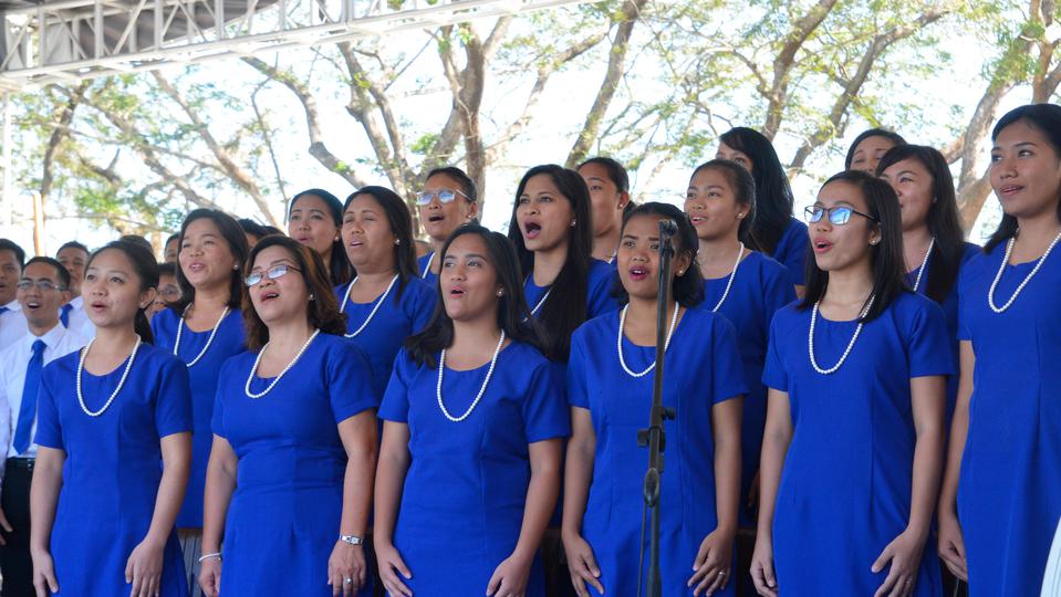 A multistake choir of female Latter-day Saints wearing blue dresses and white necklaces, singing at the Urdaneta Philippines Temple groundbreaking ceremony.