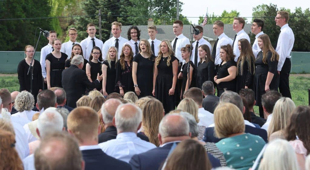 A choir of young men in the back row wearing white shirts and ties, and young women in the front row wearing black dresses, singing.