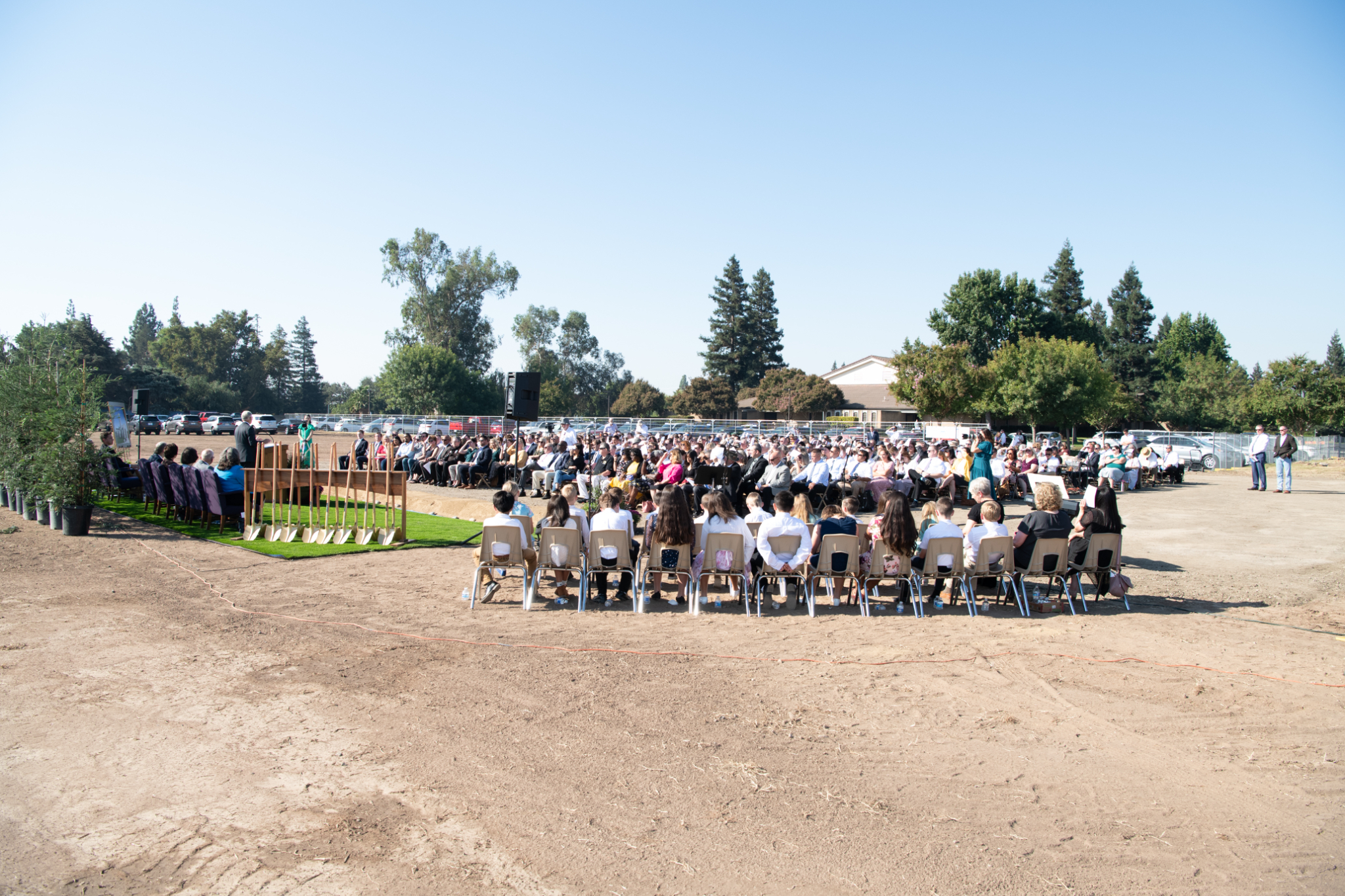 A large group of people wearing Sunday best and sitting outside in chairs on a dirt field.