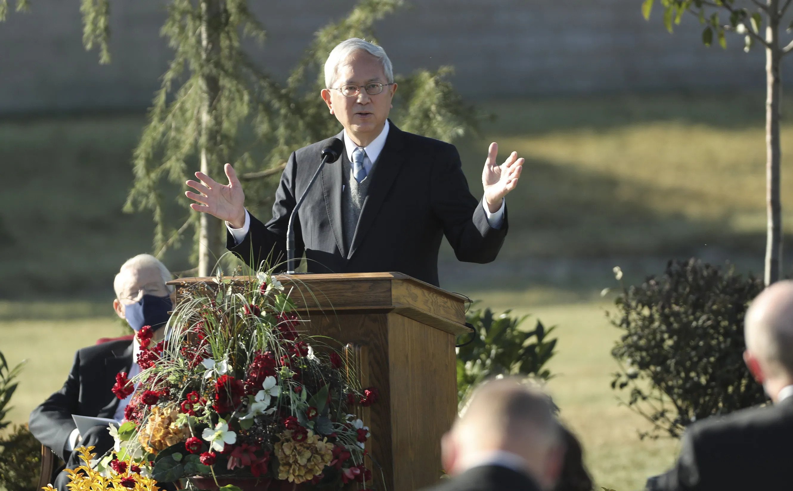 Elder Gong wearing a suit and tie and speaking from a pulpit outside with his hands pointed outward.