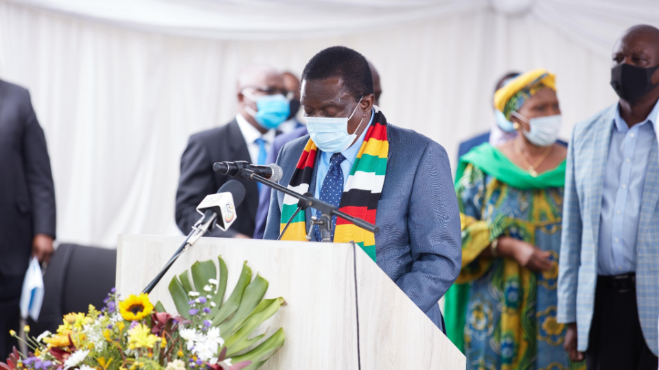 A man wearing a suit and tie, as well as a scarf with the colors of Zimbabwe's flag, speaking from a pulpit outside.