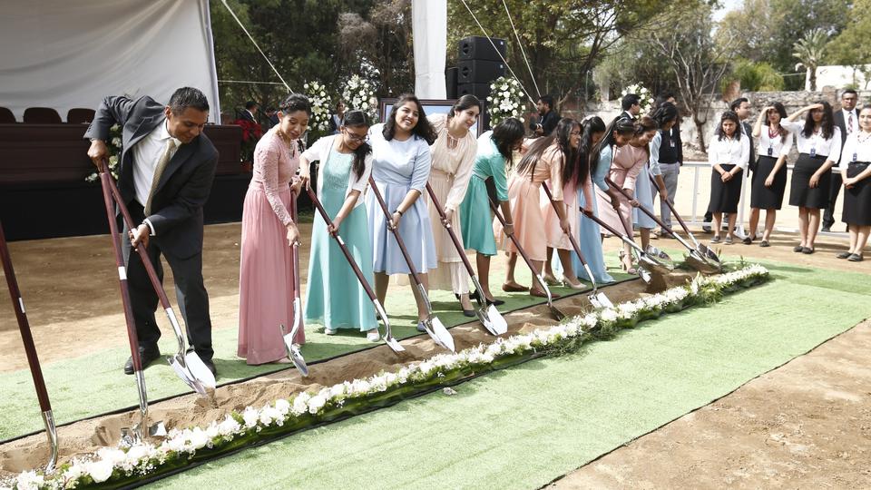 A row of young women in dresses digging into the ground with ceremonial silver shovels.