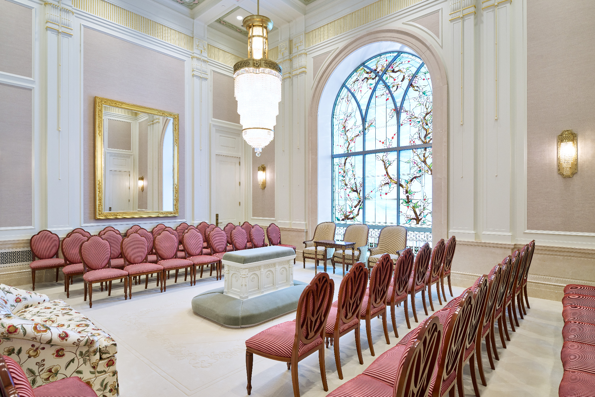A white room with an altar in the center, with rows of light-red chairs on either side and a chandelier hanging from the ceiling.