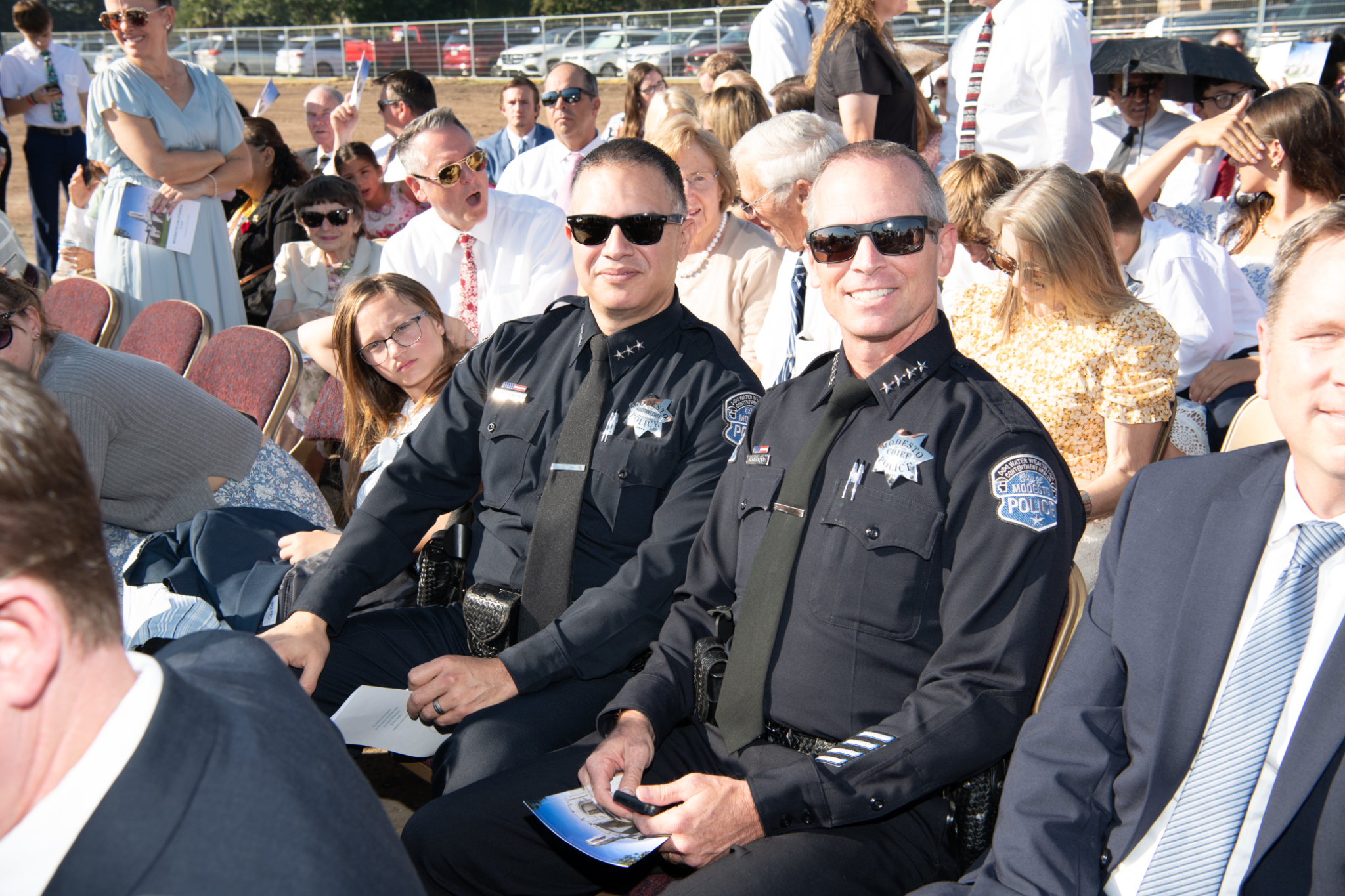 Two smiling police officers in a large group of people sitting in chairs outside.
