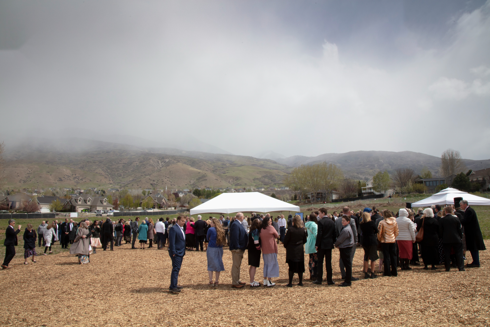 A group of people in Sunday best gathered in a dirt field, with a large grass field around them.