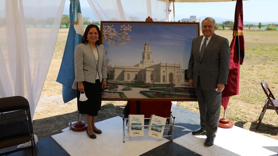 A man and a woman in Sunday best standing next to a framed picture of the rendering of the Salta temple.