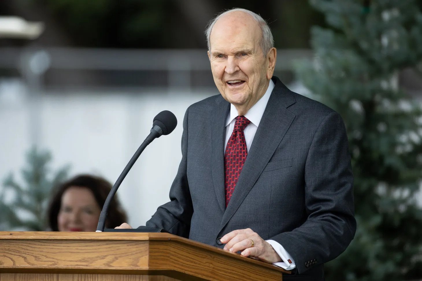 President Nelson wearing a gray suit and red tie and speaking from a pulpit outside.